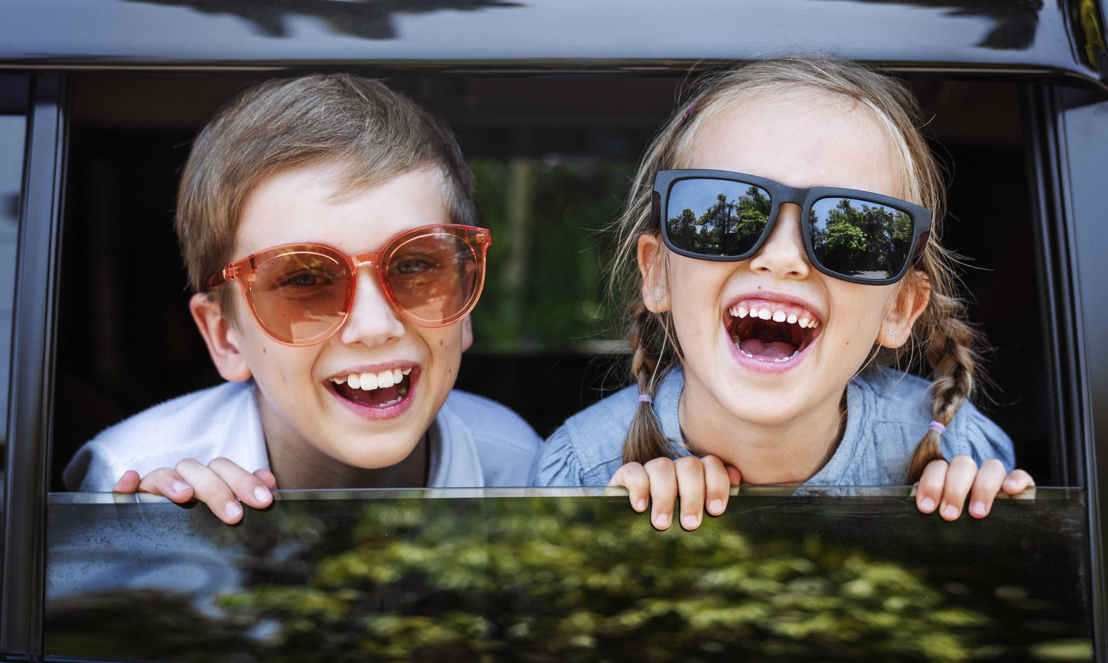 Ein Junge und ein Mädchen mit Sonnenbrillen schauen lächelnd aus dem Autofenster